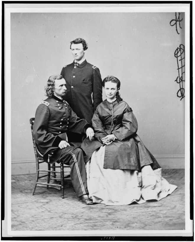 A Civil War Army general sits next to a woman in a long dress. Between them stands another man.