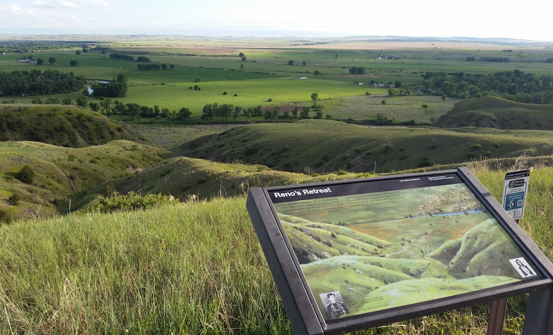 A plaque labeled “Reno’s Retreat” marks a spot at the Little Bighorn National Battlefield. In the background is a field of rolling green hills.
