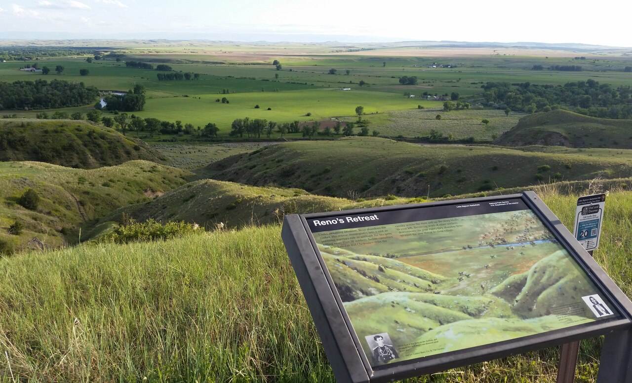 A plaque labeled “Reno’s Retreat” marks a spot at the Little Bighorn National Battlefield. In the background is a field of rolling green hills.