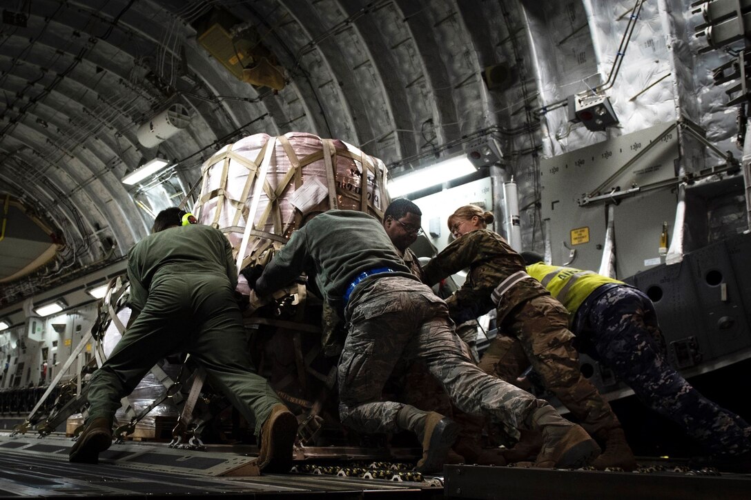 A group of service members push a heavy pallet of supplies up an aircraft ramp.