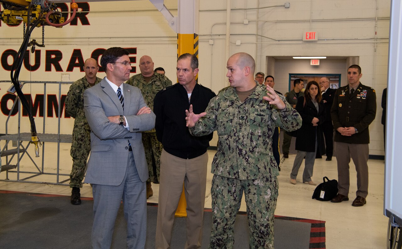 Three men stand near each other. One, in a military uniform, speaks to another.