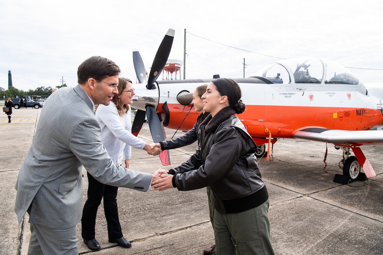 A man in a suit shakes hands with a woman in a military uniform. A military aircraft is in the background.