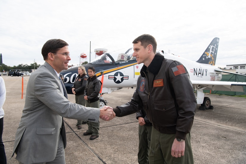 A man in a suit shakes hands with a man in a military uniform.  In the rear, a military aircraft.