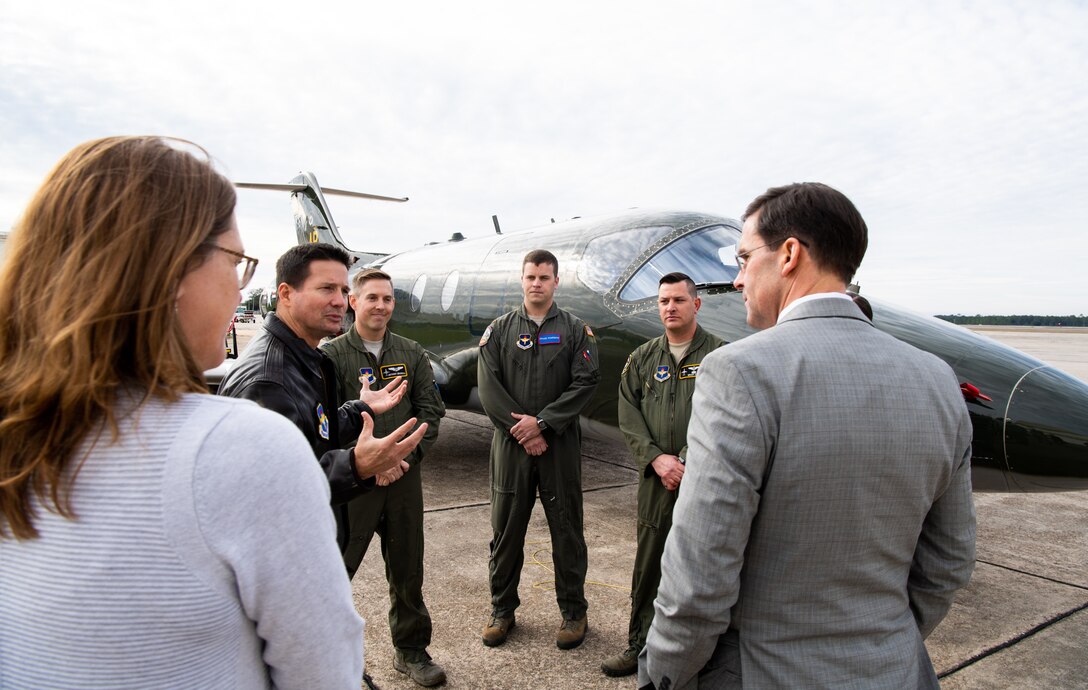 A group of people, some in military uniforms, stand in a circle and speak. A military aircraft.is in the background.