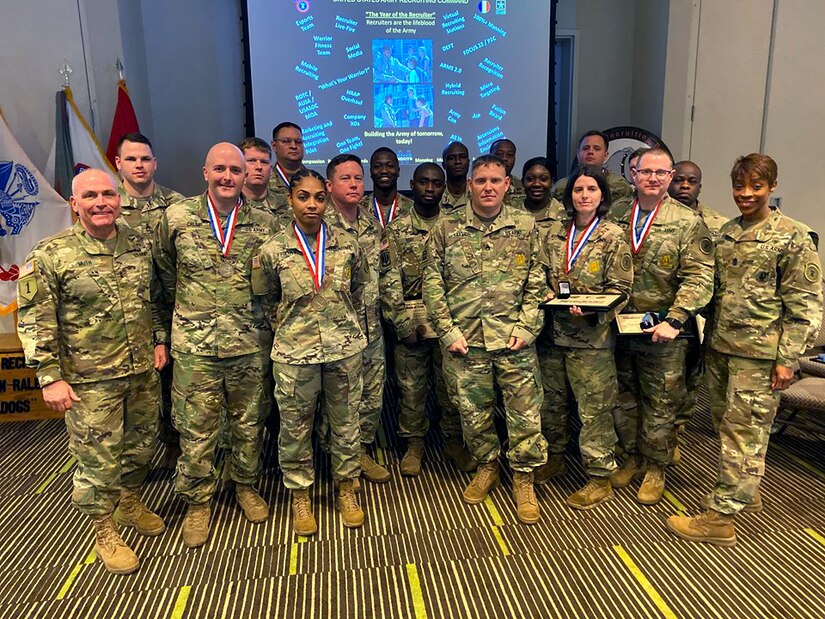 eighteen male and female soldiers in green camouflage uniforms stand in front of a screen with pictures and words on it. Some of the Soldiers have a red white and blue ribbon with a gold medallion around their necks. Three Soldiers are holding framed certificates and ring boxes. The carpet they are standing on is brown and yellow.