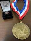 Black and white box with ring inside sitting next to a gold medal on a red white and blue ribbon sitting on a brown table.