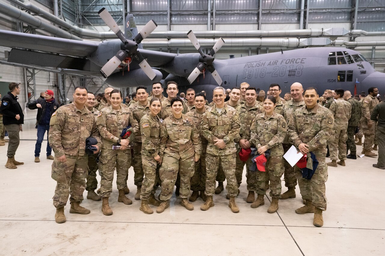 Group of men and women in military uniforms pose in front of an airplane in a hangar.