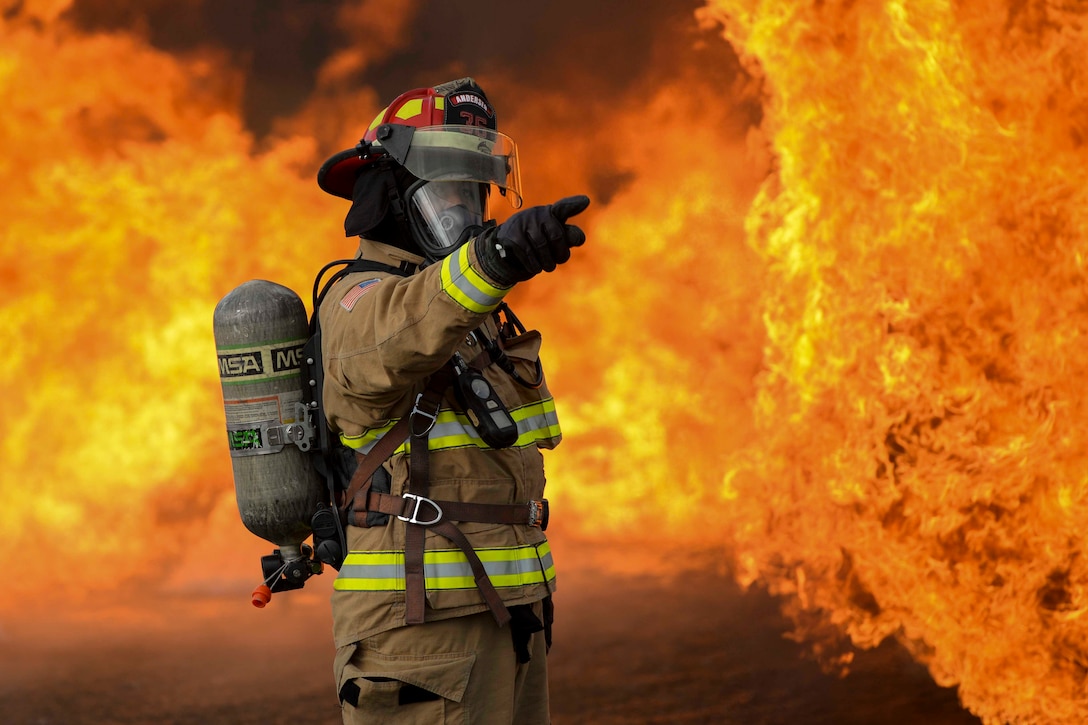 An airman points while wearing a protective suit as fire surrounds.
