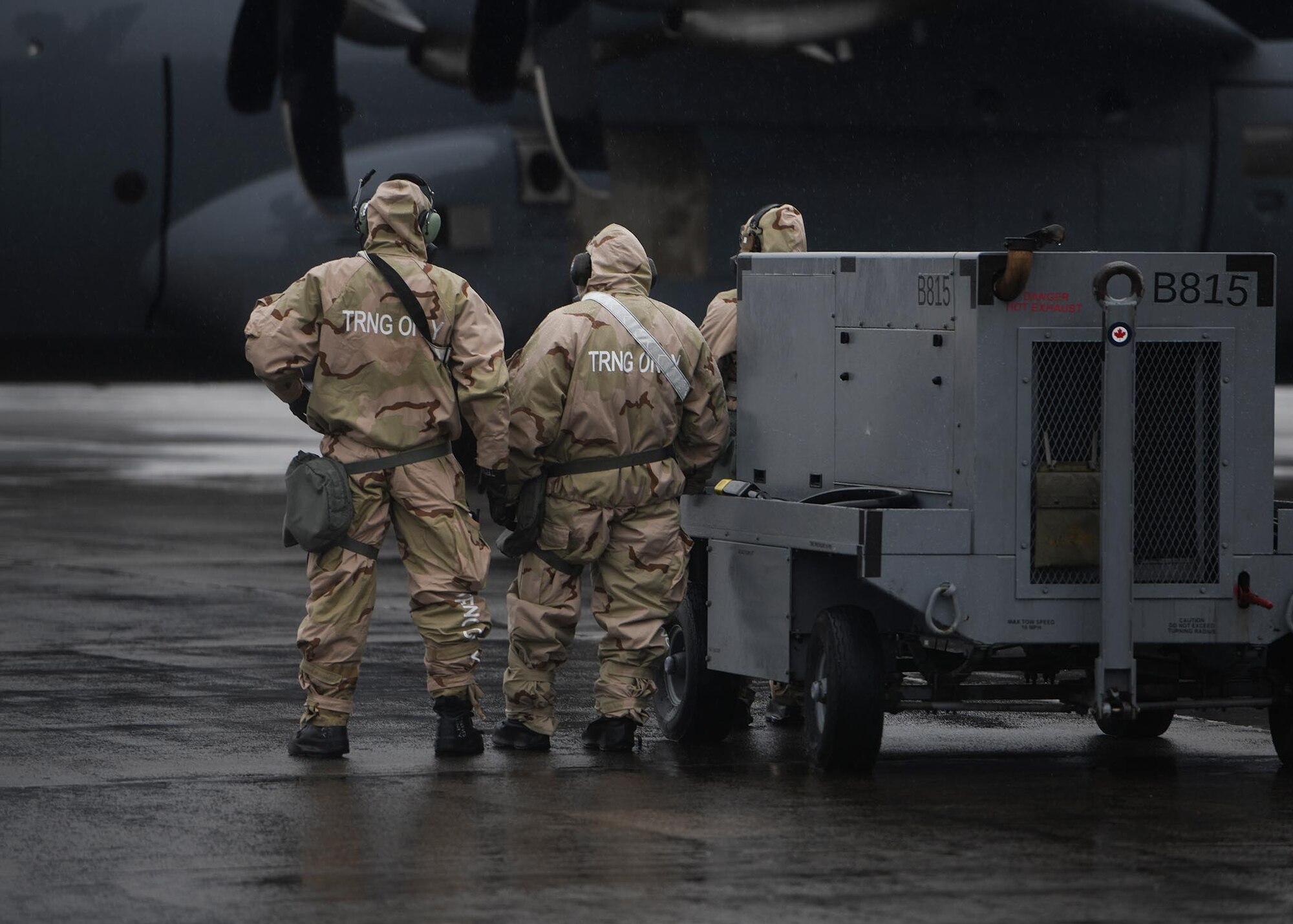 Three Airmen stand near a large grey aircraft in MOPP gear.