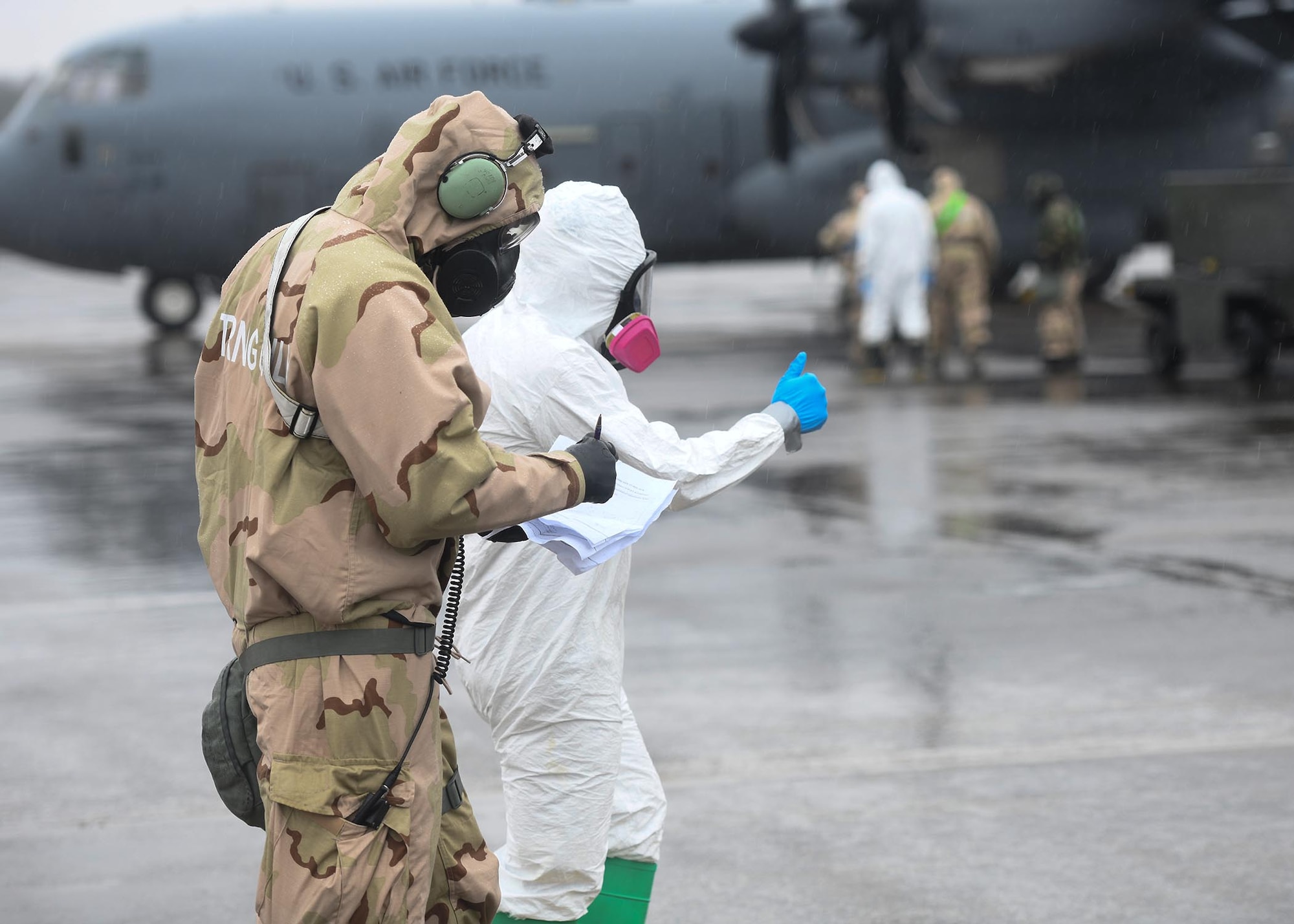 Two Airmen walk towards a large grey plane in protective clothing.