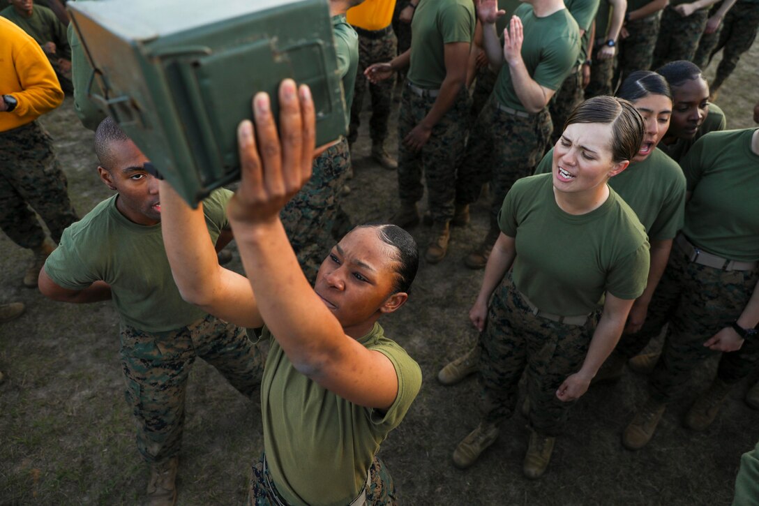 A Marine surrounded by others, lifts an ammunition can overhead.