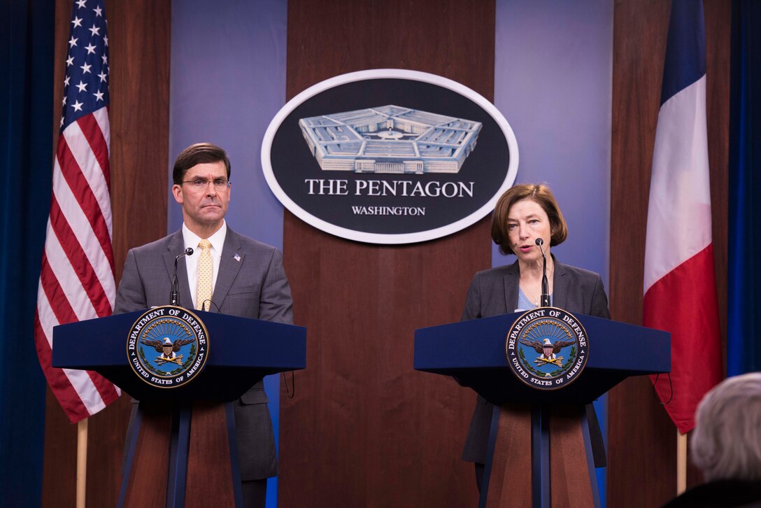 Two people stand at podiums in front of a Pentagon sign and an American flag and a French flag.