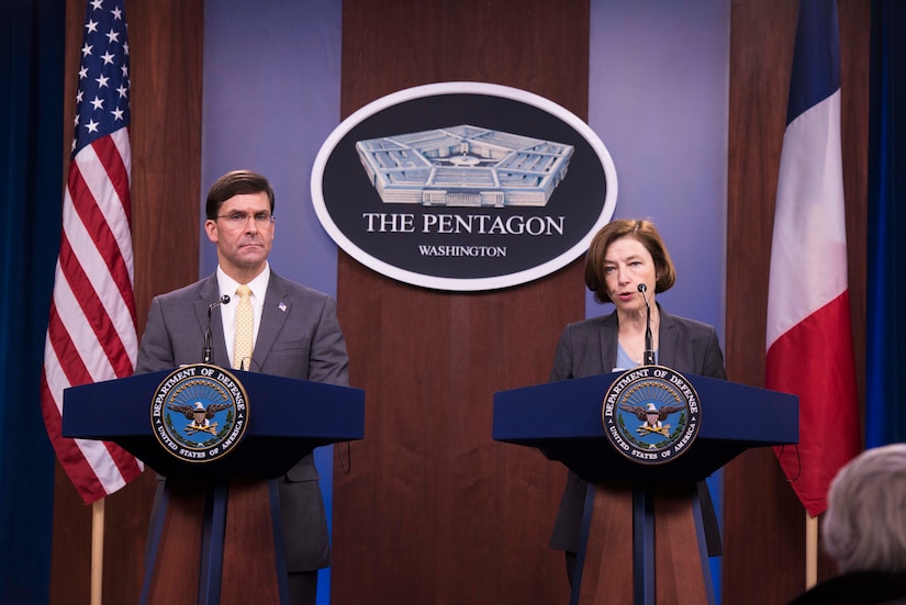 Two people stand at podiums in front of a Pentagon sign and an American flag and a French flag.