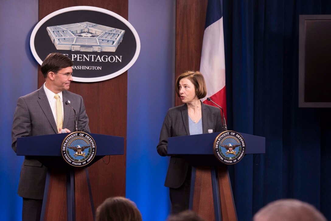 Two people stand at podiums in front of a Pentagon sign.