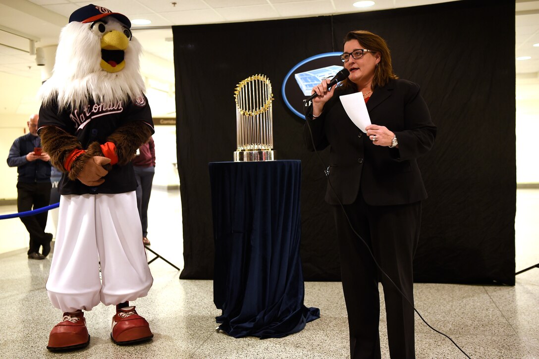 A woman holding a microphone stands in front of a trophy. Nearby is a person in a large bird costume.