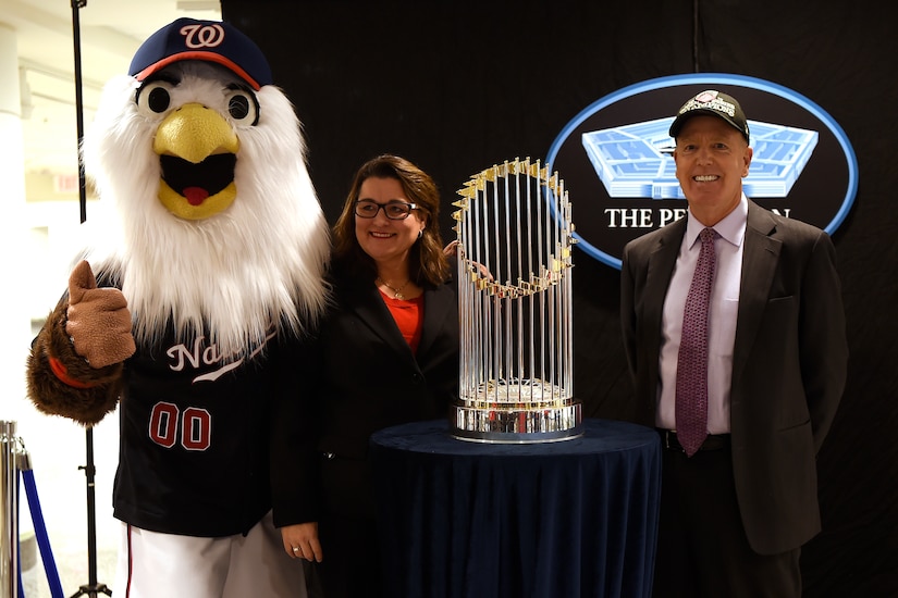 A person in a large bird costume, a woman and man stand behind a trophy, which is situated on a small table.