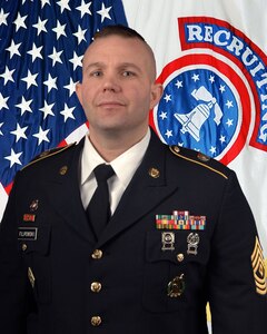 White male Soldier in Army dress blue uniform jacket, white shirt and tie, with colorful ribbons on the right and first sergeant rank on his sleeve, standing in front of an American flag and a recruiting flag.