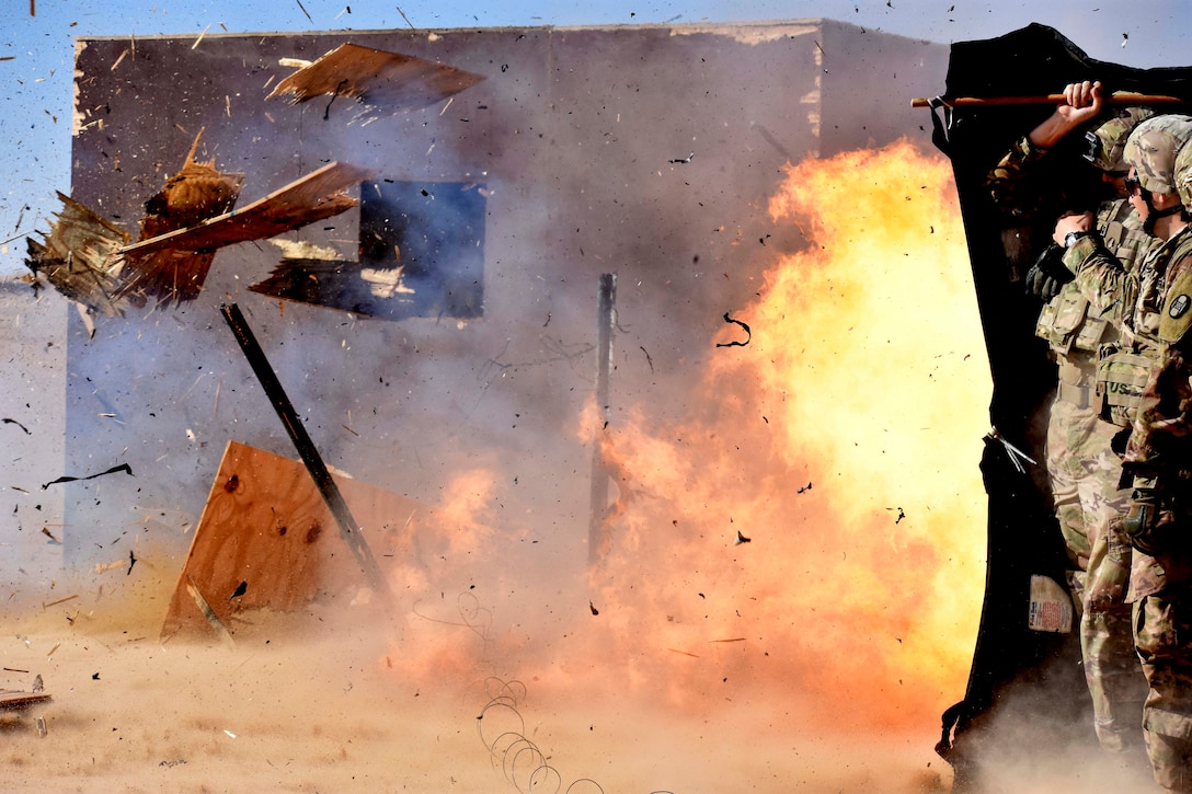 Soldiers stand behind a barrier as an explosive detonates.