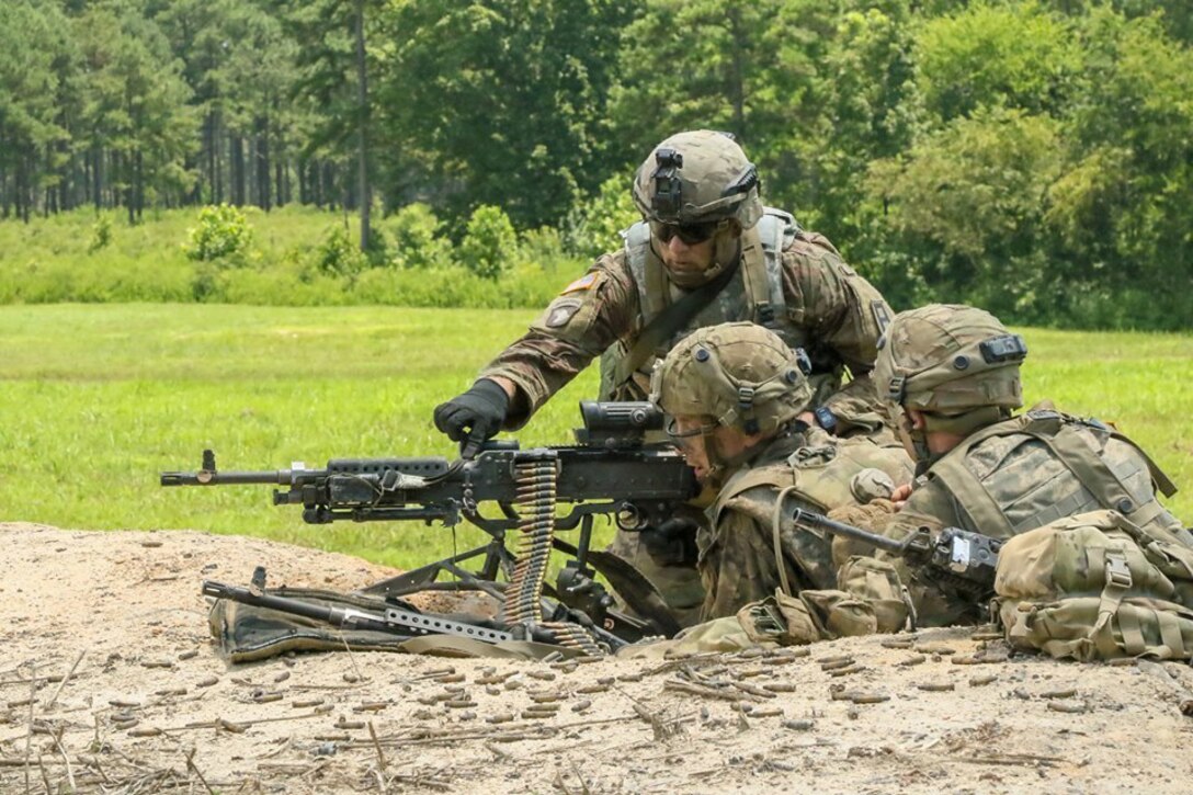 A soldier points to a machine gun as two other soldiers lie on the ground and listen.