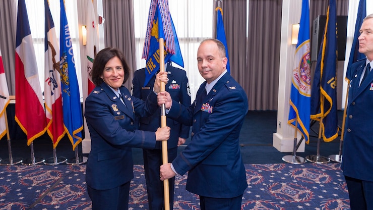 Two people, a woman on the left and a man on the right, in dark blue Air Force service dress uniforms are both holding a guidon flag. Another man in the dark blue Air Force service dress uniform is standing at the position of attention on the right side of the picture. There are numerous, multicolored flags in the background.