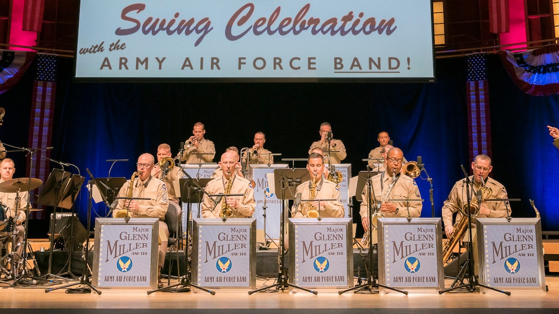 About 15 instrumentalist are sitting and performing while dressed in the light brown uniforms that look like what was worn during Army Air Force days. Above them on the large screen are the words: Swing Celebration, with the Army Air Force Band. There are five signs in front of the front row of musicians that read: Major Glenn Miller and his Army Air Force Band.
