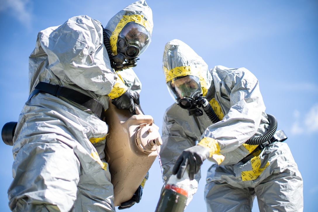 Two men wearing protective clothing pour a liquid onto a simulated chemical weapon.