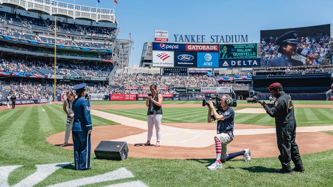 Woman in dark blue Air Force ceremonial uniform singing the national anthem from behind home plate in Yankee Stadium. Four people are standing and kneeling a few feet in front of her with cameras in their hands, and the words "Yankee Stadium" is visible in the distance with several multicolored advertisements below, and a large screen can be seen projecting the image of the vocalists face.