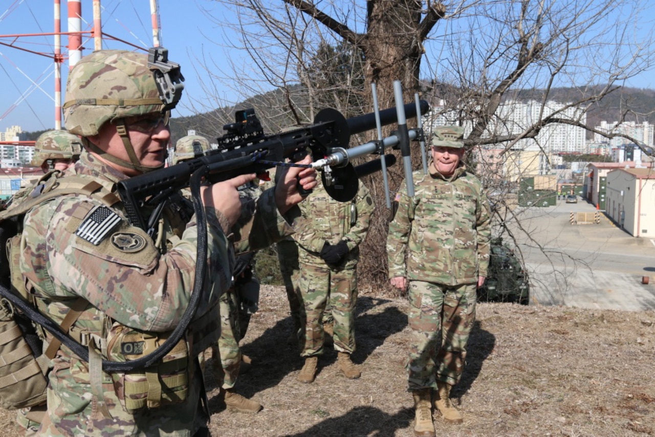 Men in military uniforms stand outside watching a soldier holding a rifle.