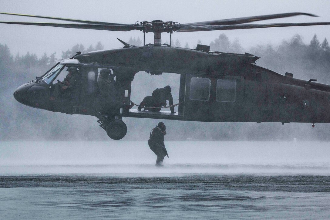 A soldier stands outside of a helicopter in the rain while other soldiers sit inside the aircraft.