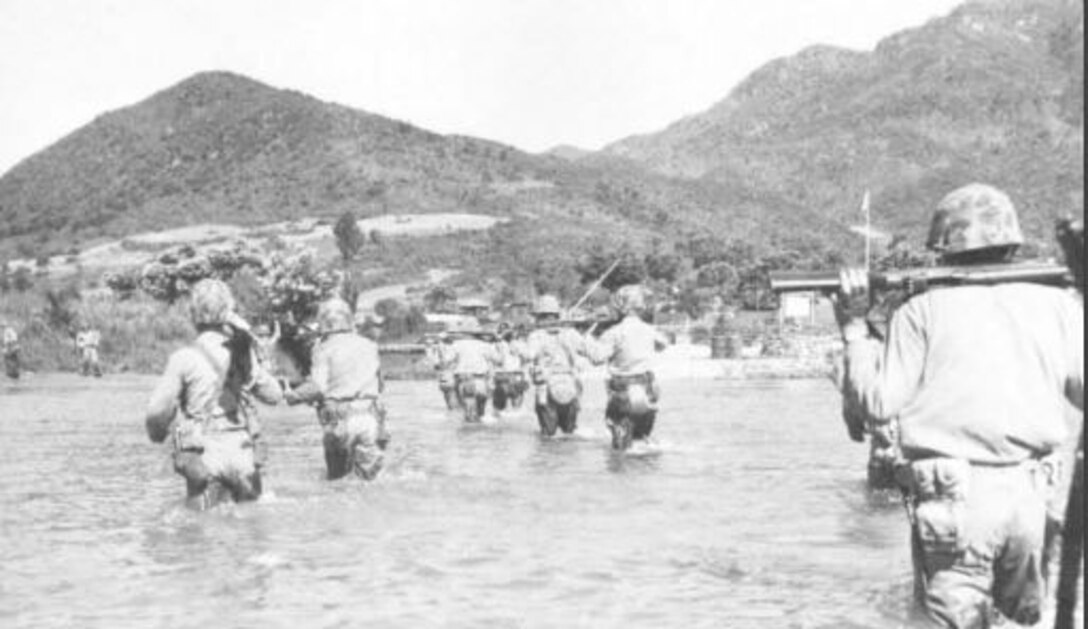 Several Marines wade through thigh-high water carrying weapons on their backs, walking toward a hill in the distance.