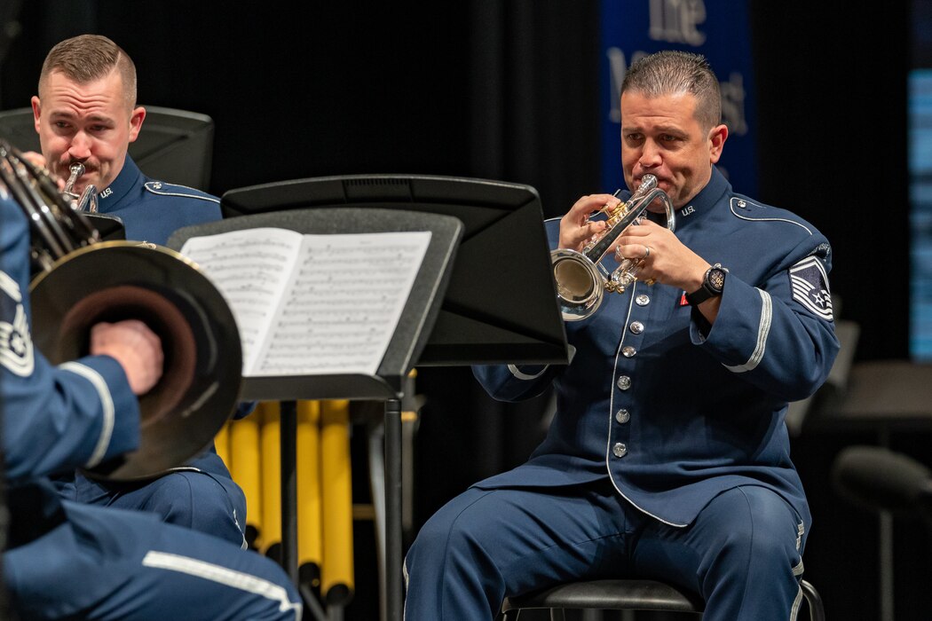 Two male trumpet players are seated while playing their instruments and are dressed in the dark blue Air Force ceremonial uniform.