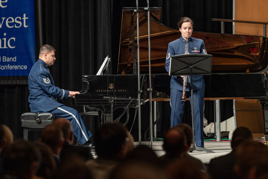 A female English horn player performs in front of a male pianist, who is sitting and performing on a black grand piano. Both are dressed in the dark blue Air Force ceremonial uniform. Behind them and to the left is a blue banner with white text that is partially obscured. The backs of the heads of dozens of audience members is seen in the foreground, the bottom portion of the picture.
