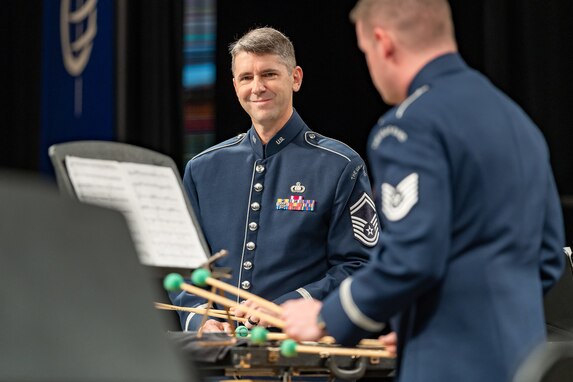 Two percussionists are standing while playing their instruments and are dressed in the dark blue Air Force ceremonial uniform. The musician in the middle of the picture is smiling. They are performing with mallets that have bright green balls on the ends of tan colored sticks.