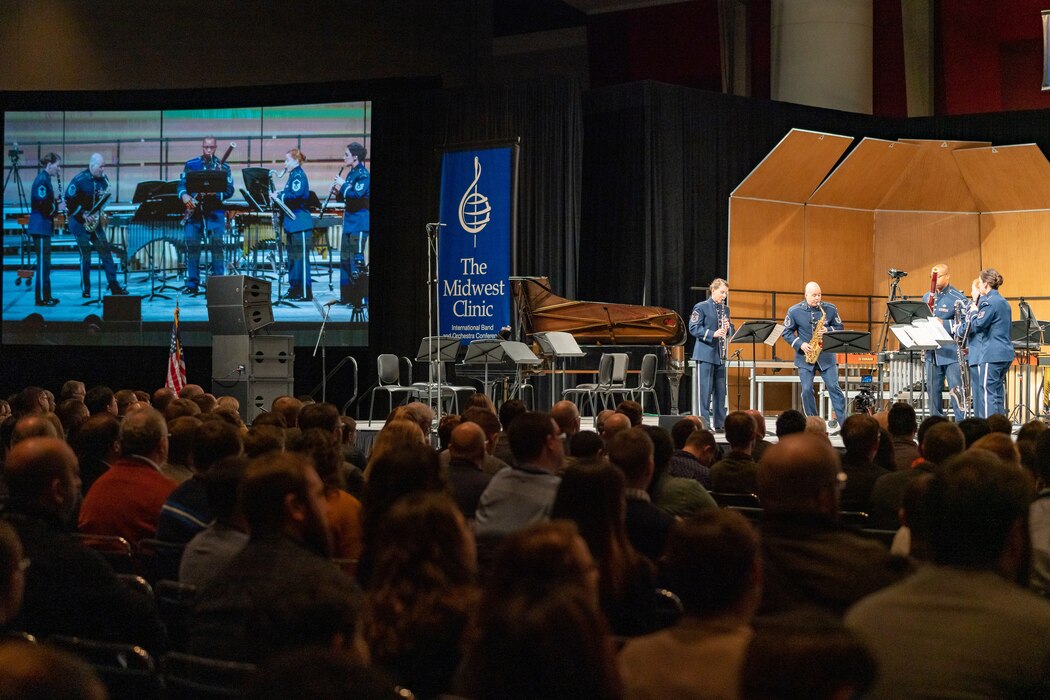 Five musicians are seen both on stage and on a large screen to the left. They are standing while playing their instruments and are dressed in the dark blue Air Force ceremonial uniform. There is a blue banner in the middle of the picture which reads "The Midwest Clinic," and the backs of the heads of dozens of audience members is seen in the foreground, the bottom portion of the picture.