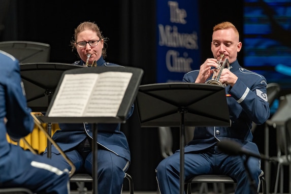 Two trumpet players, a male and female, are performing seated on stage dressed in the dark blue Air Force ceremonial uniform. The words "The Midwest Clinic" is on a banner in the background.