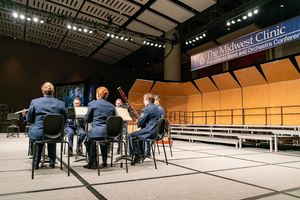 Six musicians are seated and performing various woodwind instruments on stage dressed in the dark blue Air Force ceremonial uniform. Above the stage, to the right side, is a banner which reads "The Midwest Clinic, International Band and Orchestra Clinic."