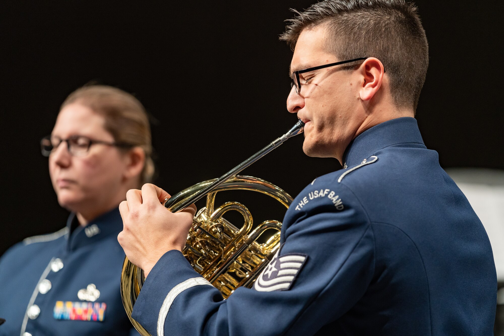 Man performing the French horn while wearing the blue Air Force ceremonial band uniform, with a woman in the background.