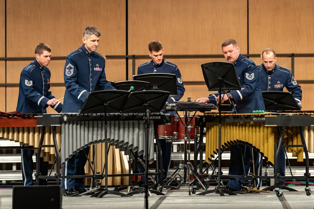 Five Air Force Band members dressed in the dark blue Air Force ceremonial uniform are playing percussion instruments on a stage with a light brown background.