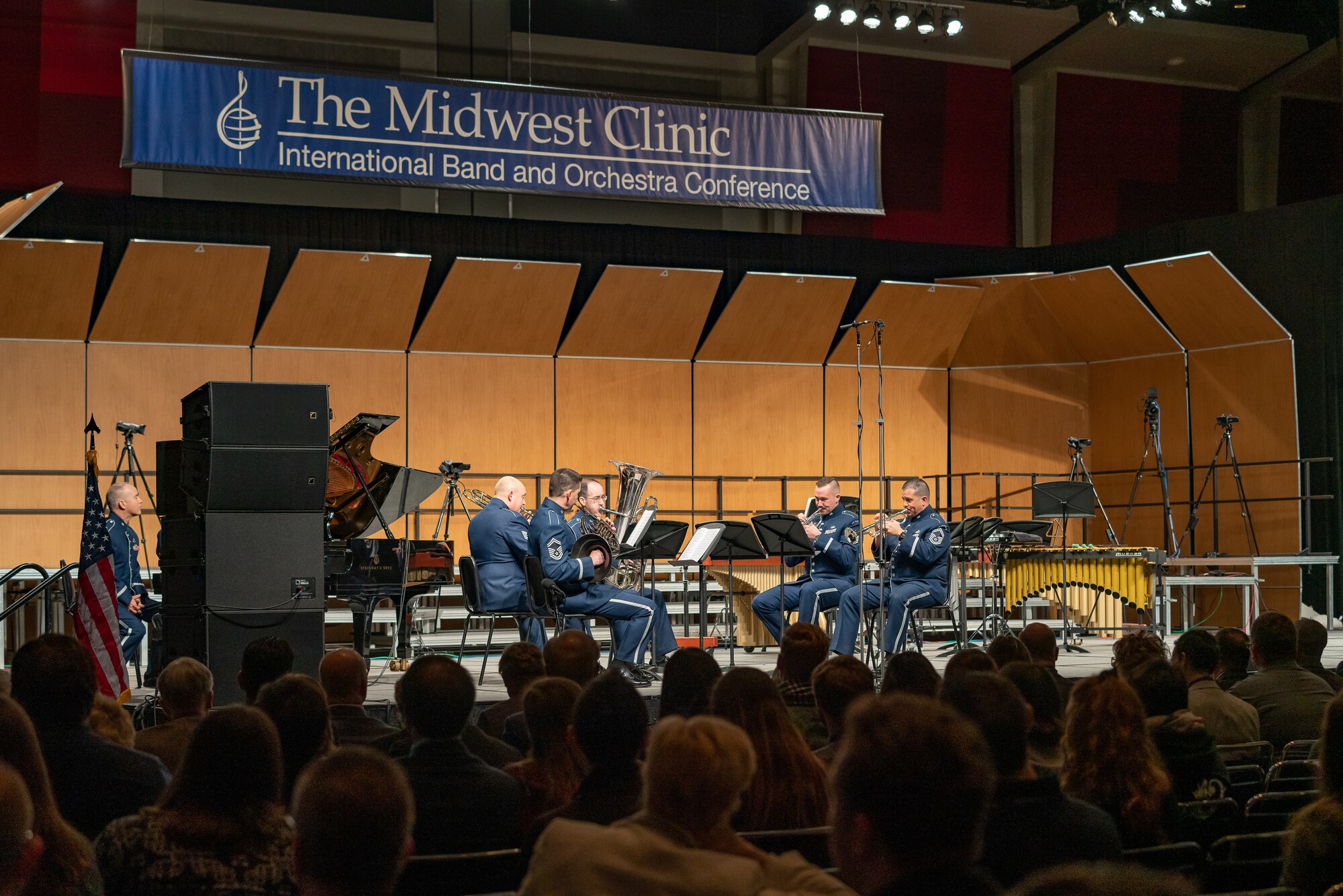 Instrumental musicians wearing the blue Air Force ceremonial uniform performing on a stage with a blue banner above which reads: The Midwest Clinic, International Band and Orchestra Conference. The backs of the heads of dozens of audience members is seen in the foreground, the bottom portion of the picture.