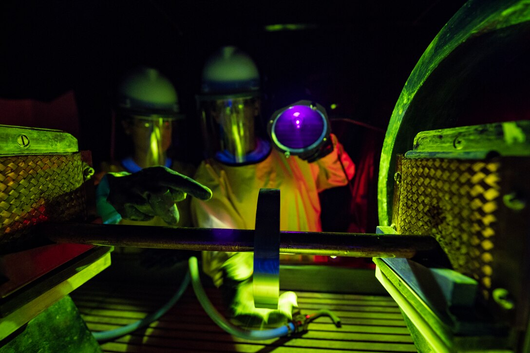 Two airmen look at equipment wearing protective gear.