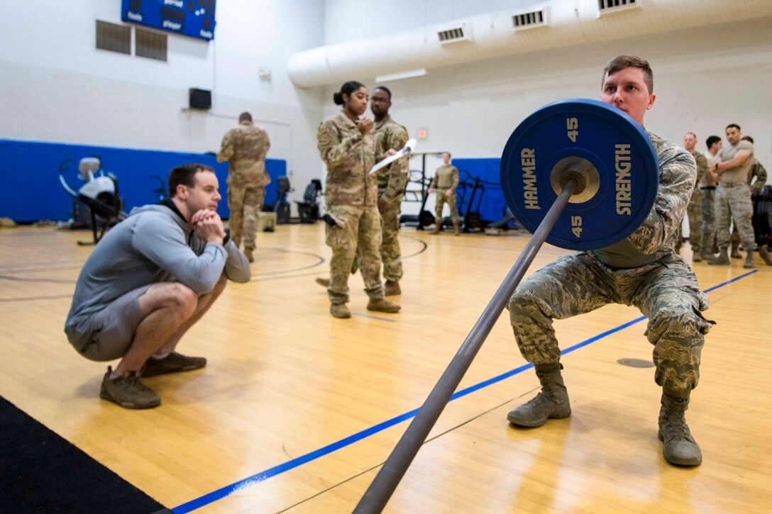 An airman squats down while holding a weightlifting bar; others surround.