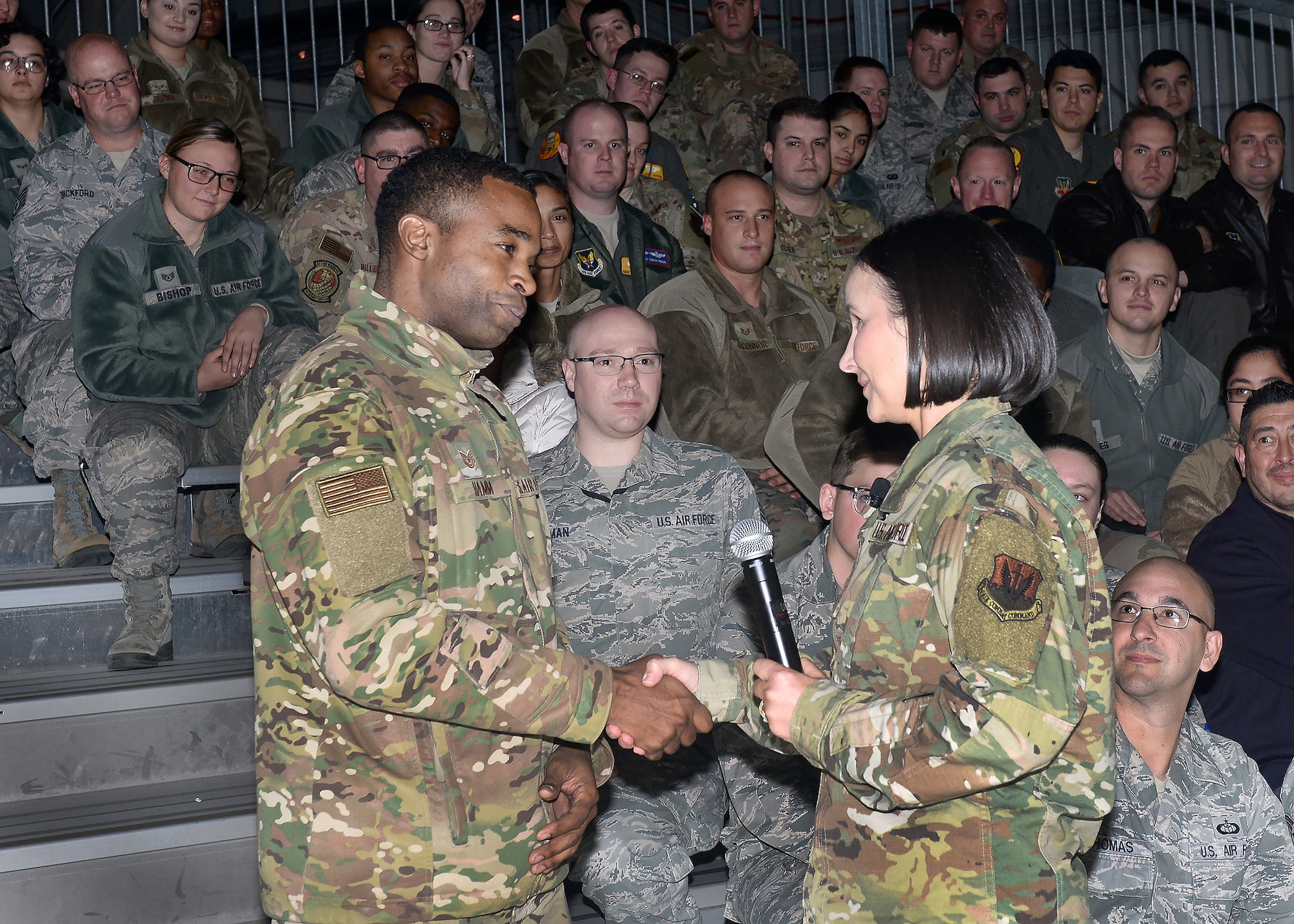 with an audience in the background, 16th air force command chief Leifer is shaking Technical Sergeant Hamm's hand with large audience in the background