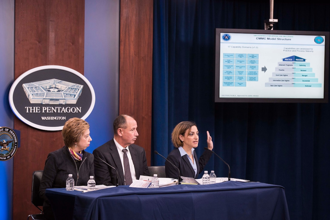 Three people sit behind a desk and talk to reporters at a briefing.