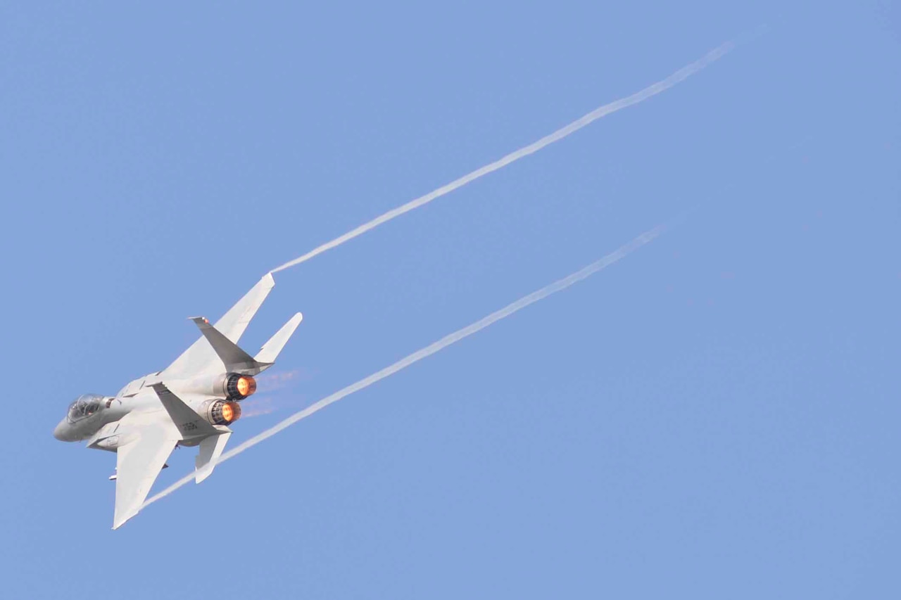 An F-15 Eagle fighter jet flies across a blue sky with jet stream tailing behind it.