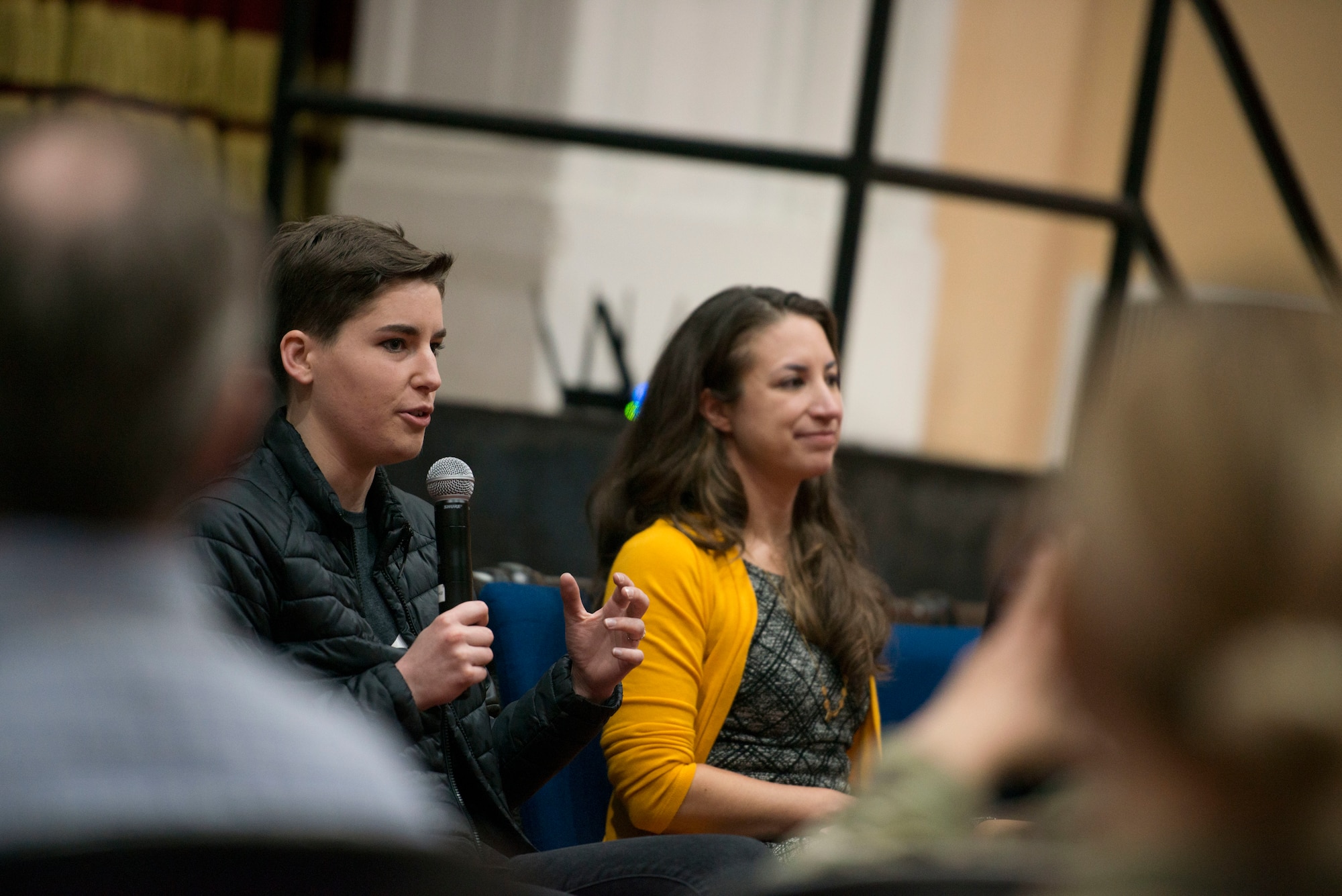 A photograph of a woman holding a microphone and speaking to an audience