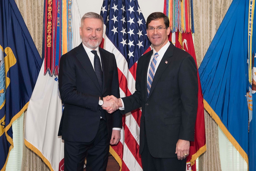 Two men standing in front of flags shake hands.