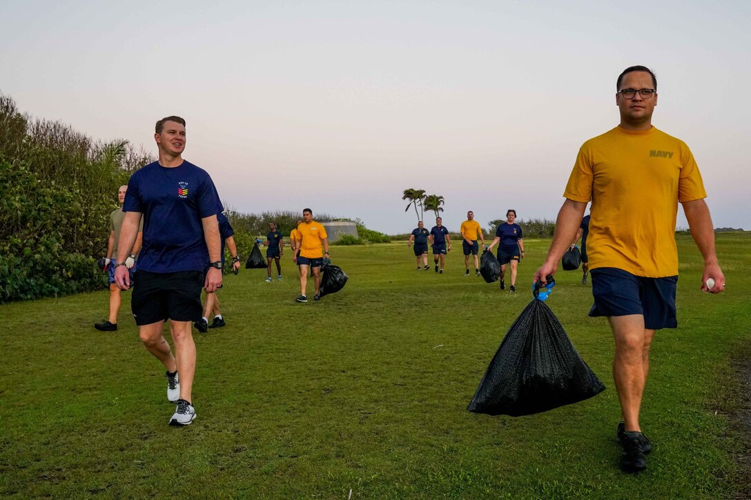 Sailors carry trash bags on a grassy shoreline.