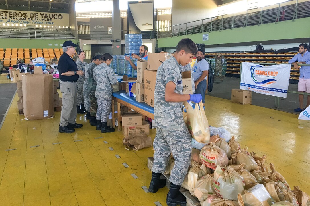 Service members handle bags of supplies in a large room.