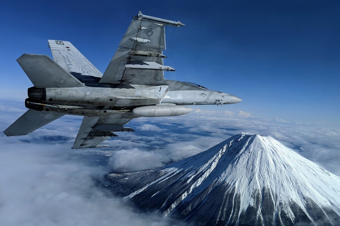 A military jet flies above the clouds near a beautiful mountain.