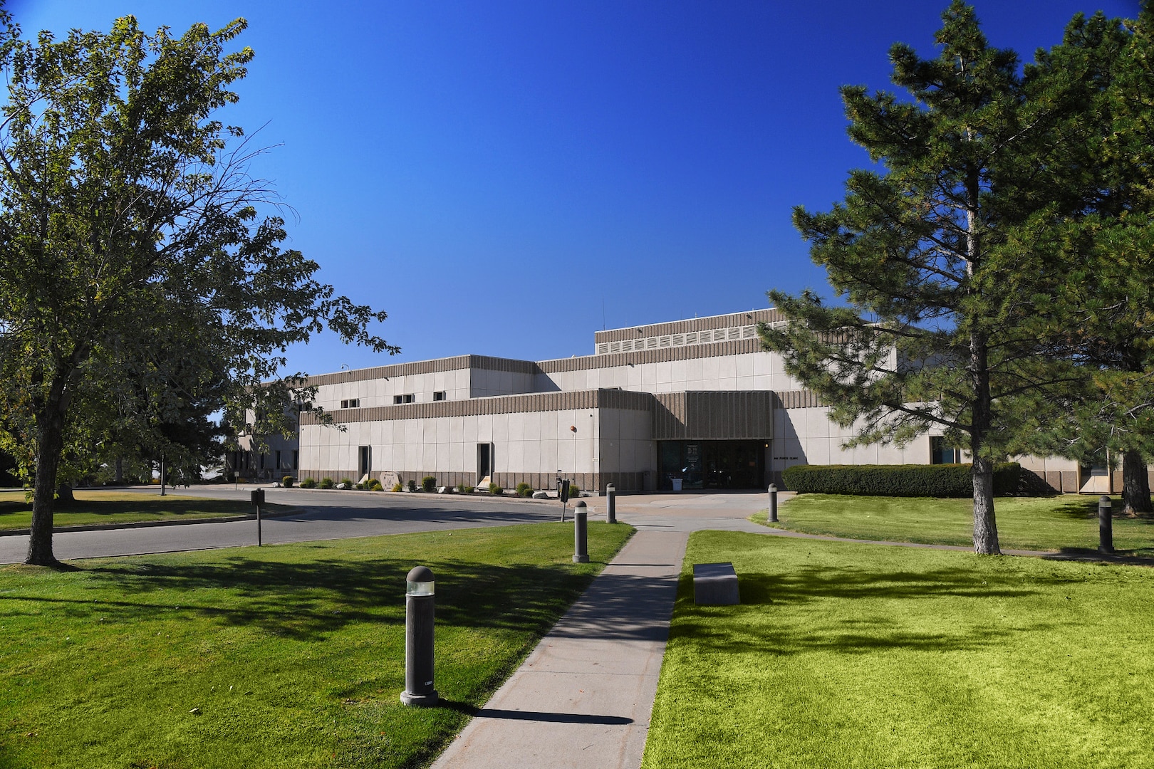 An image of the 75th Medical Group Military Treatment Facility. A cloudless, blue sky is seen over the top of the building that is flanked by trees on the left and right. Green grass and a sidewalk leading up to the MTF is in the foreground.